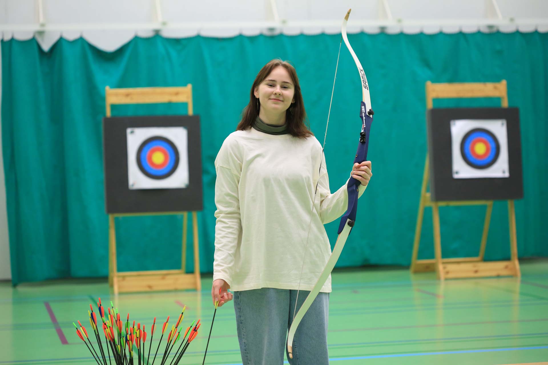 Kuvassa kasvatus- ja ohjausalan opiskelija Rianne Särkioja poseeraa jousipyssy kädessään jumppasalissa. 